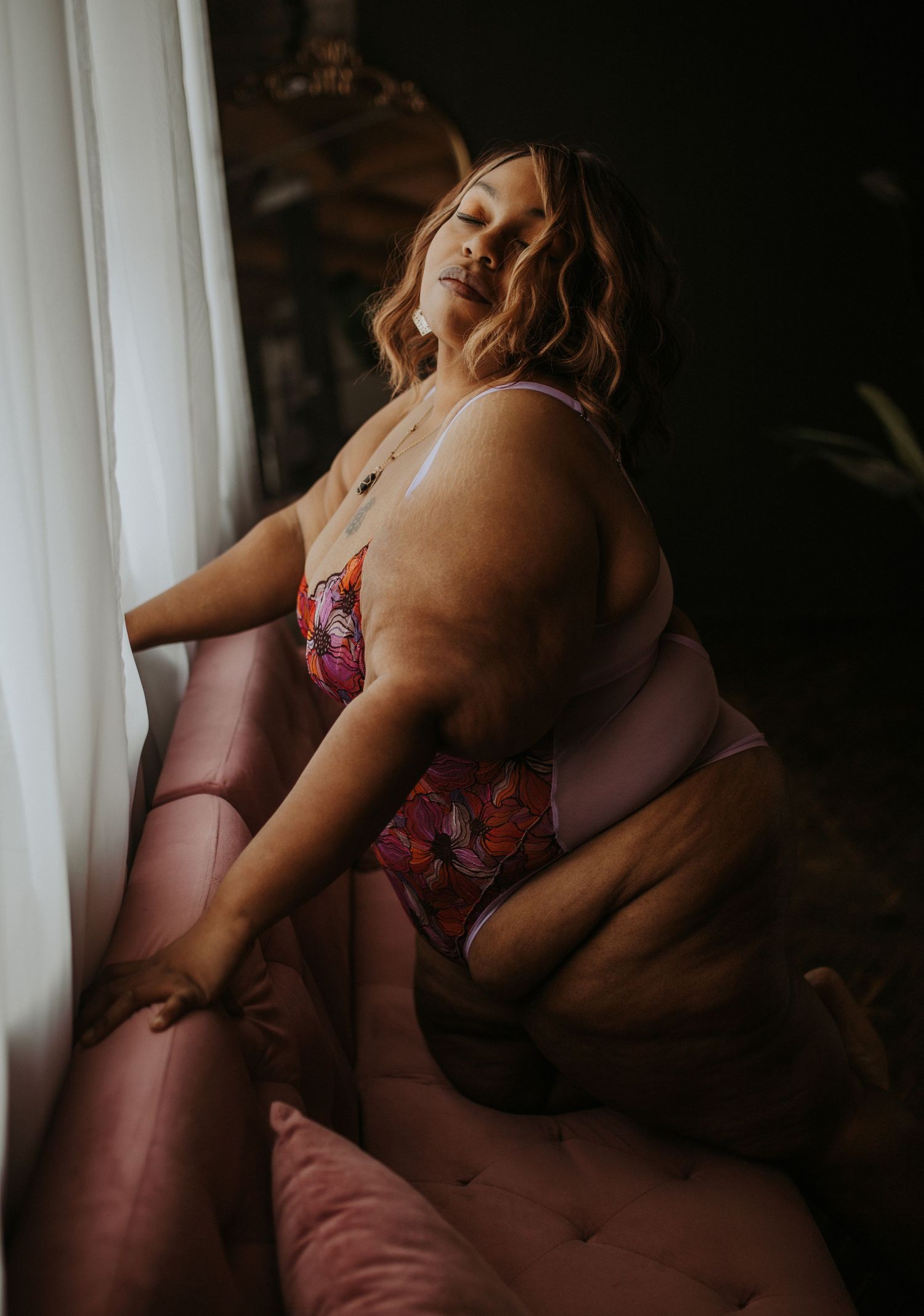 A plus-size woman poses confidently in colorful floral lingerie near a window with soft natural light.