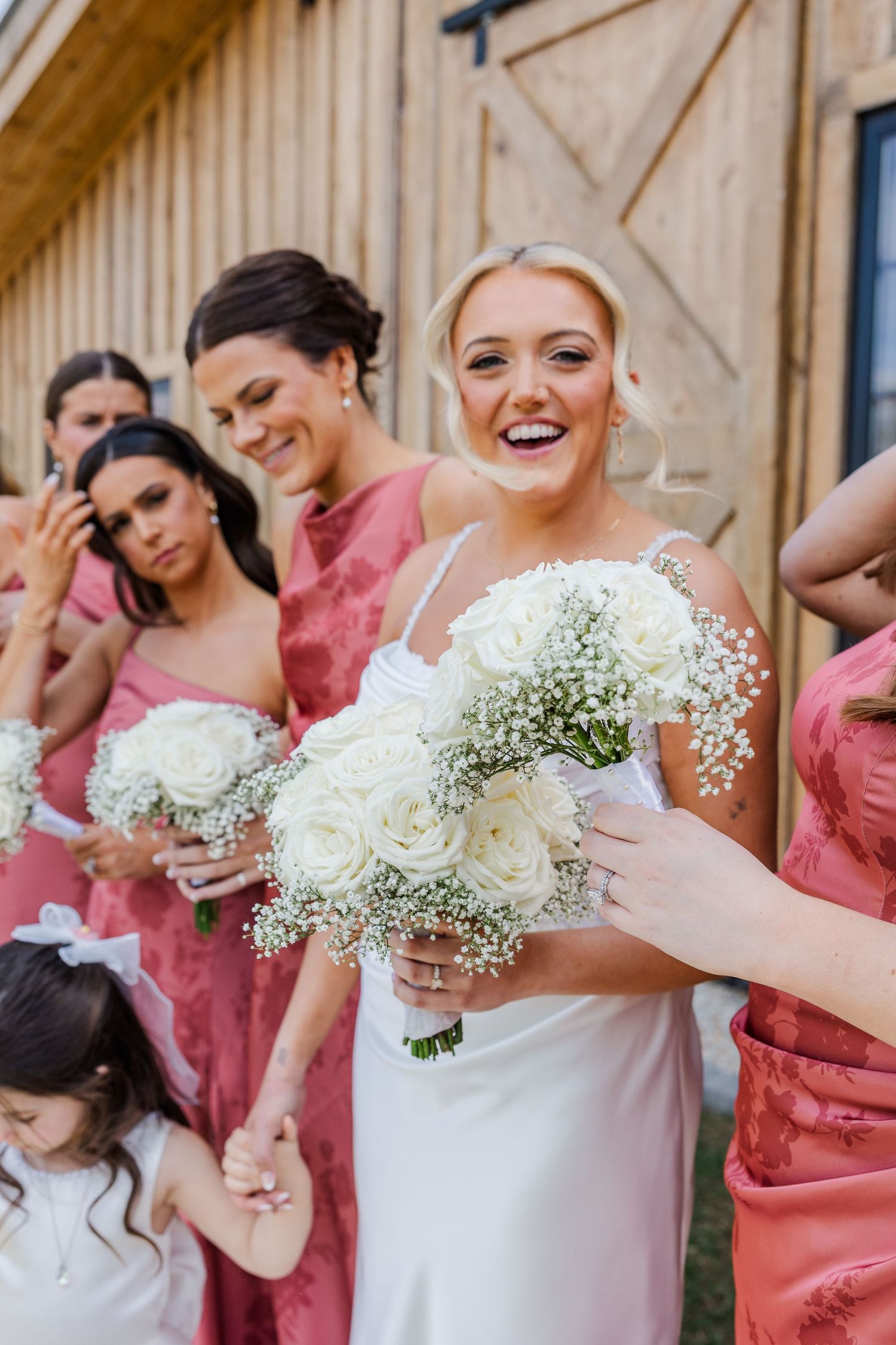Smiling bride in white gown holds bouquet of white roses surrounded by bridesmaids in pink dresses near a wooden barn.