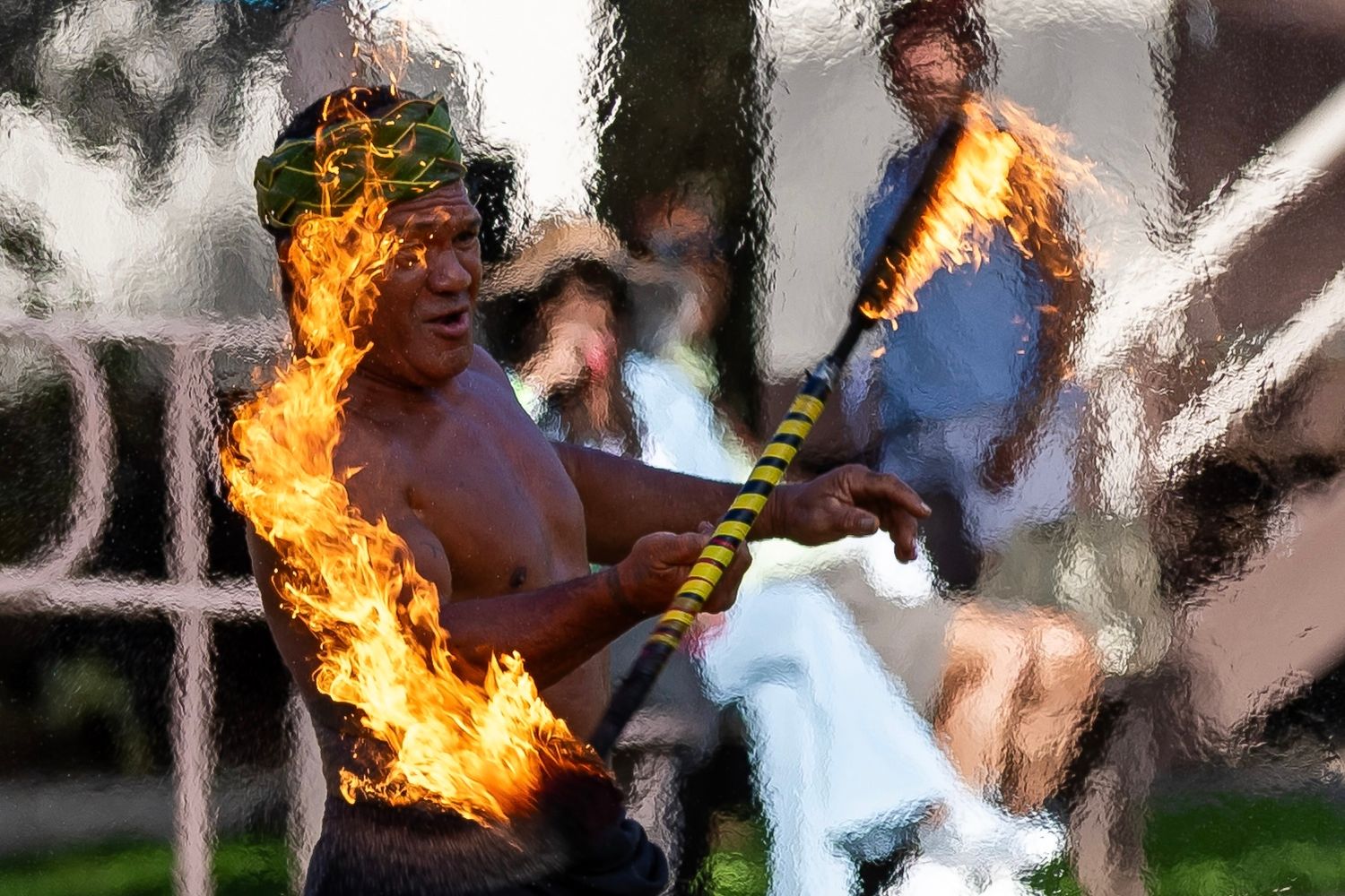A shirtless fire dancer performs a traditional Polynesian fire knife dance outdoors, wielding a flaming baton.