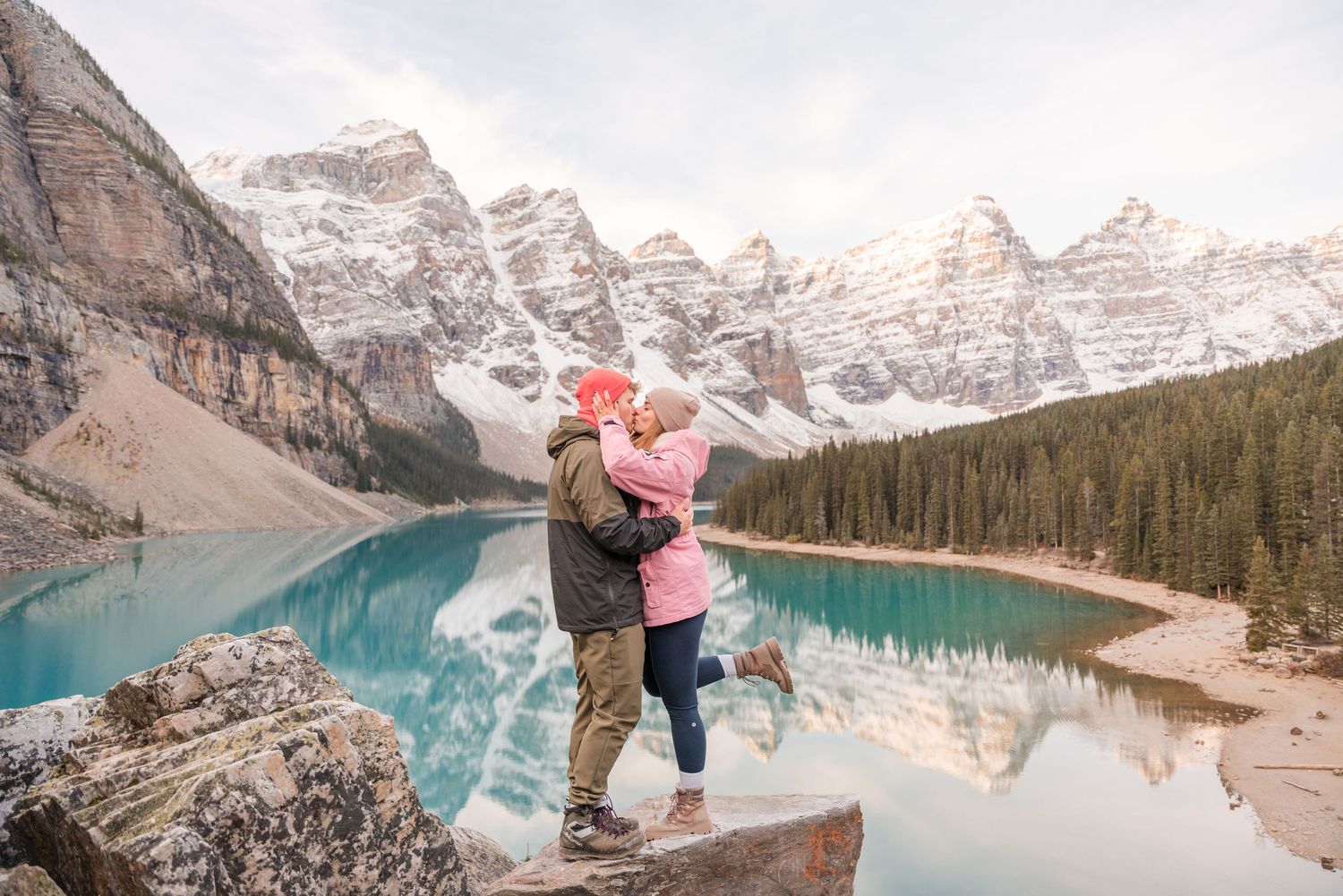 Man proposes to woman on one knee at scenic Moraine Lake, Banff, with mountains and evergreens in background.