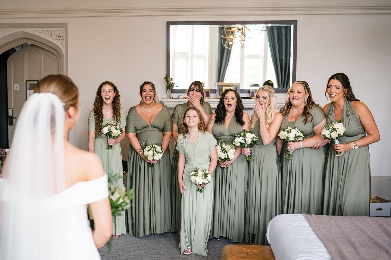 Bridesmaids in sage green dresses react with joy and surprise as bride reveals her wedding look in elegant room.