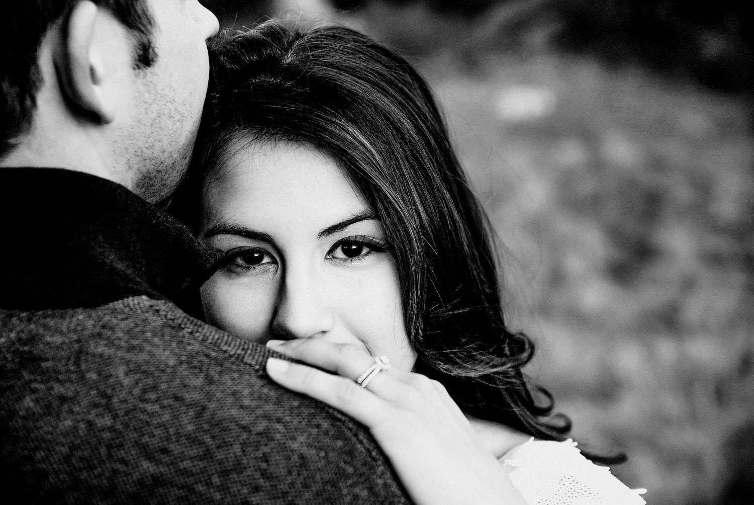 Black and white photo of a couple embracing, woman with dark hair looking over mans shoulder wearing an engagement ring.