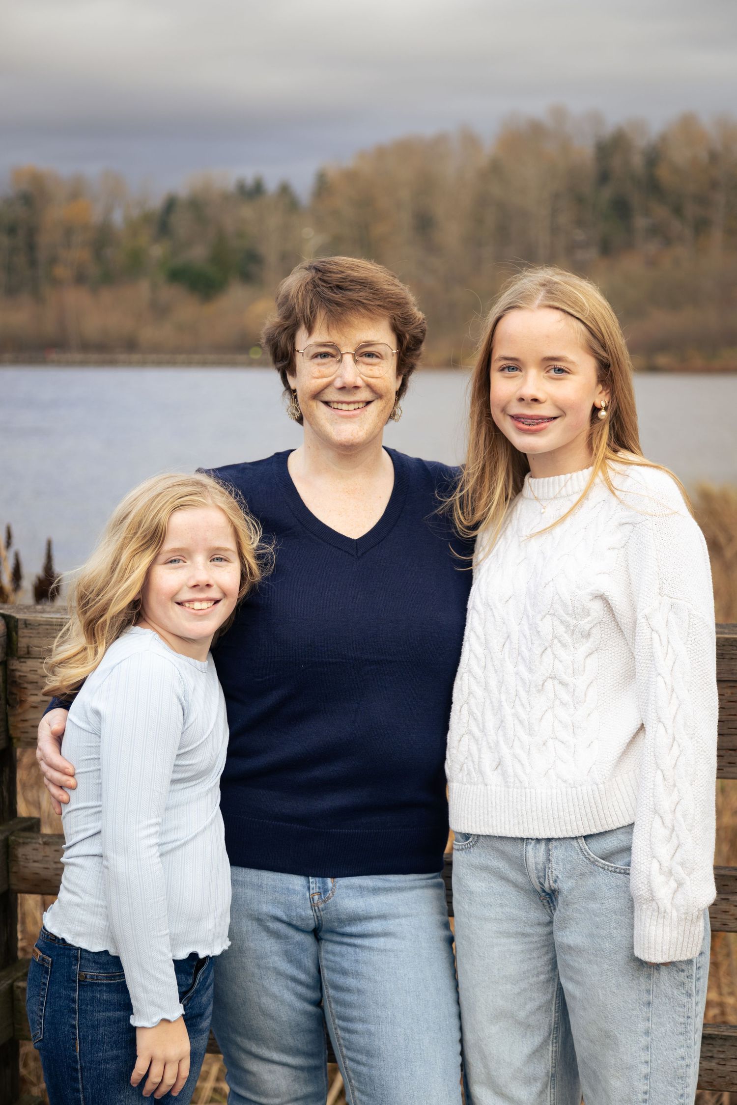 A woman and two blonde girls smile together outdoors near a lake with autumn trees in the background.