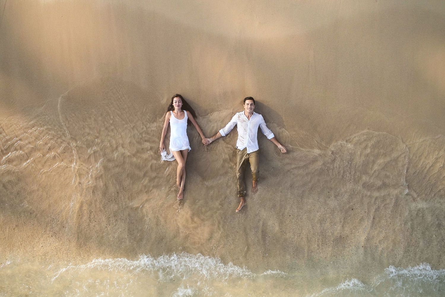 Aerial view of a couple holding hands on a sandy beach with gentle ocean waves washing ashore.