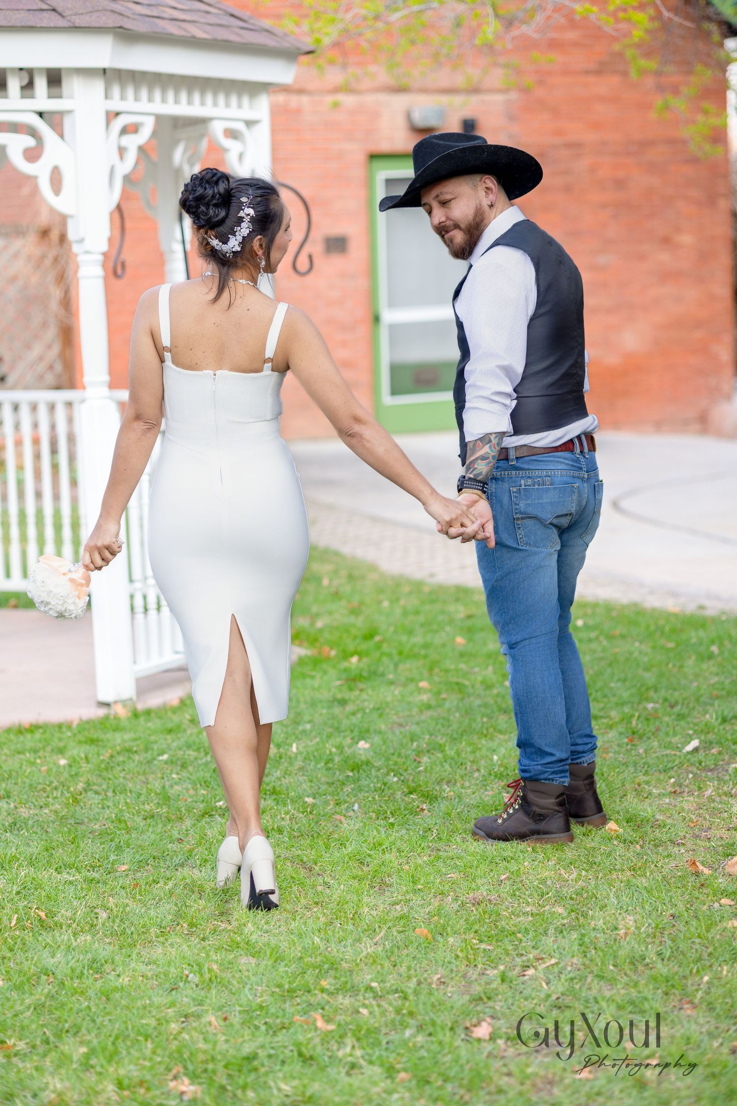 A couple holds hands in their western-inspired formal attire while walking on grass near a brick building.
