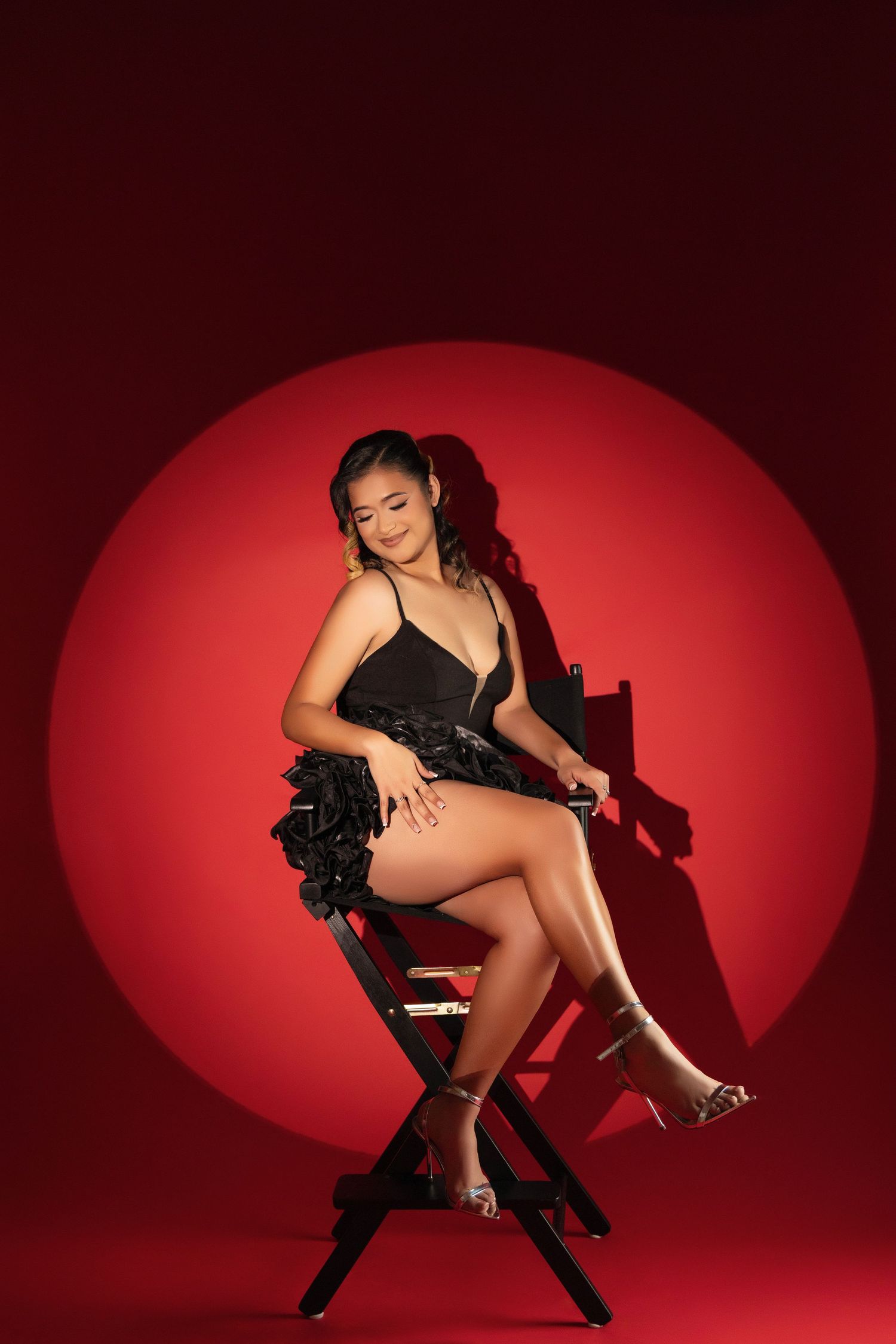 Woman in black dress poses on director's chair against dramatic red circular backdrop.