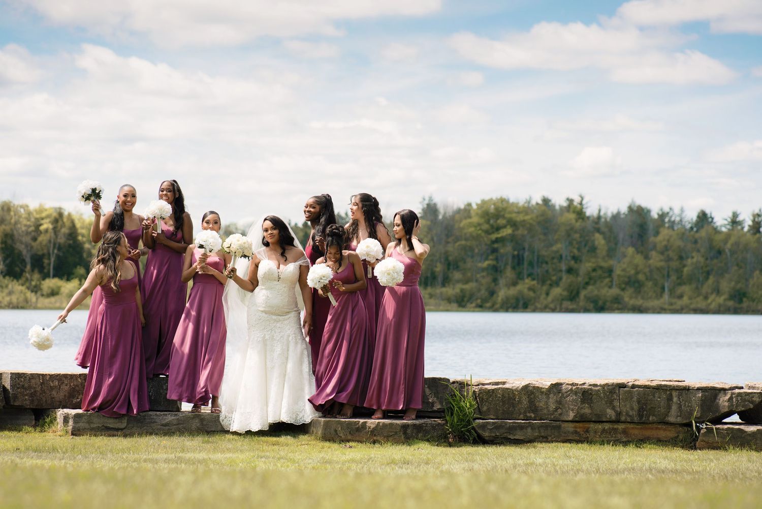 A wedding party in purple bridesmaid dresses and white gown pose together by a serene lakeside on a sunny day.