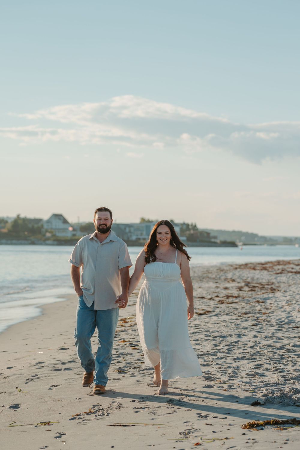A couple walks together along a scenic beach at sunset with houses visible in the background on a peaceful evening.