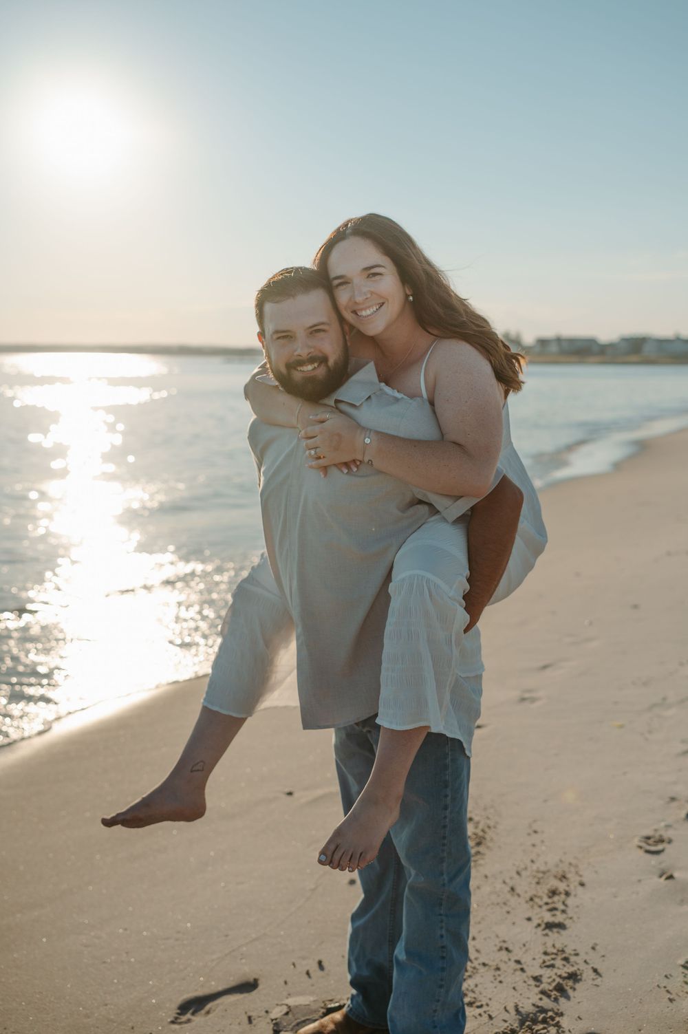 A joyful couple sharing a piggyback ride on a sunny beach with sparkling water in the background.