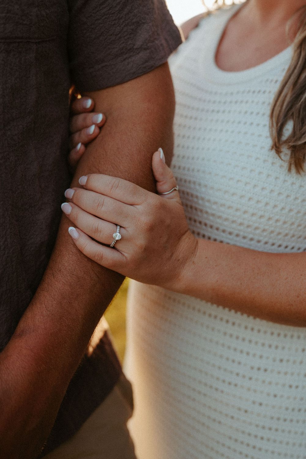 Close-up shot of hands wearing engagement ring with solitaire diamond on white crochet dress background.