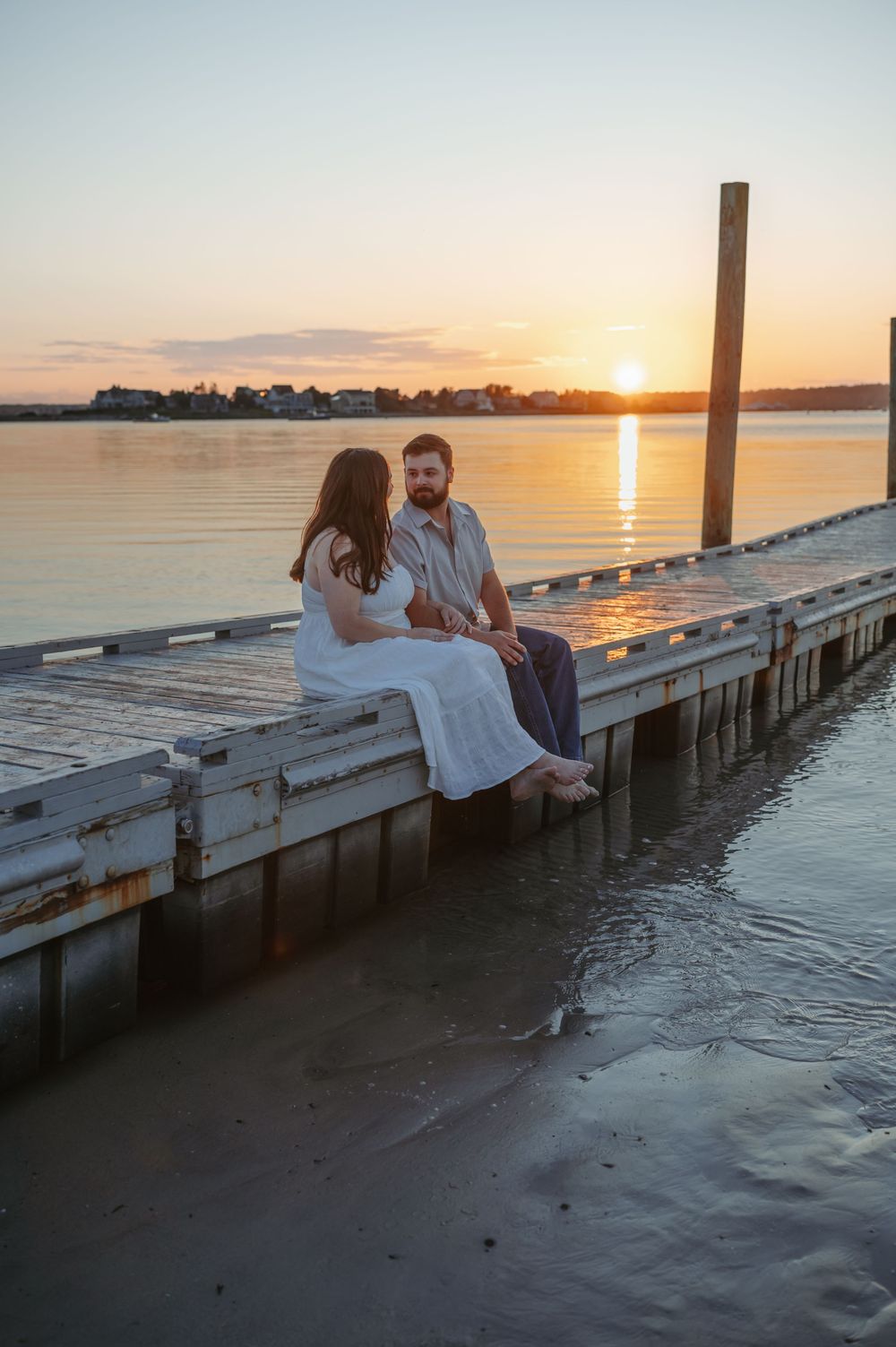 A couple sits together on a wooden dock at sunset over calm waters, creating a romantic silhouette against the golden sky.