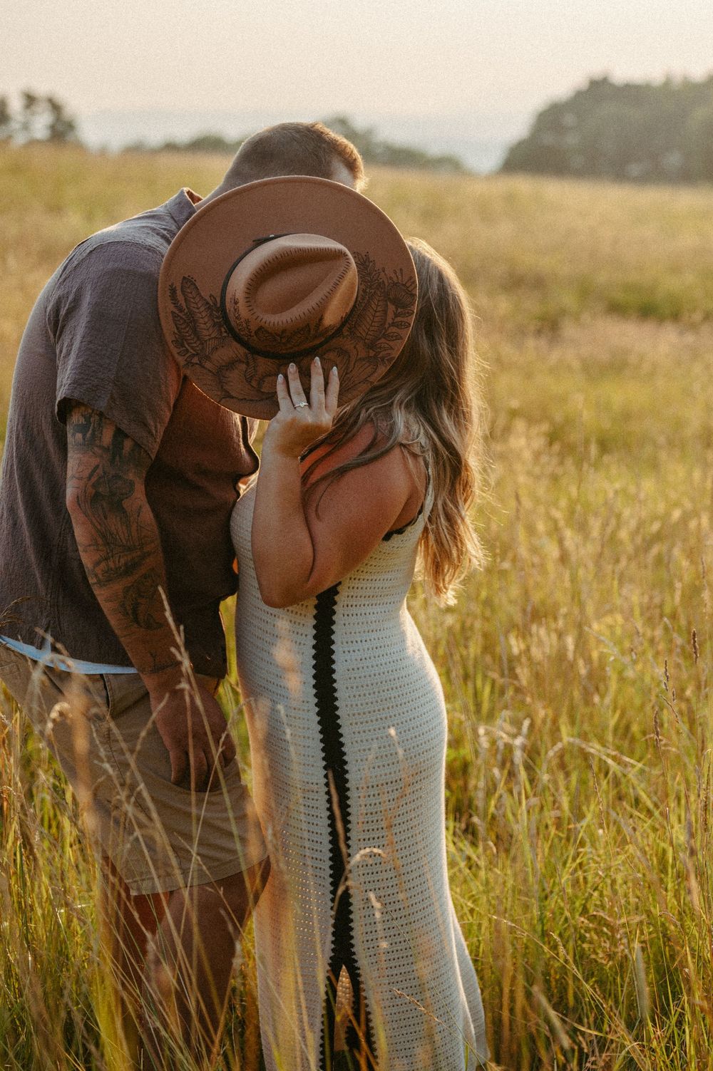 A romantic couple embraces in a sunlit field of tall grass during golden hour, with a brown cowboy hat visible.