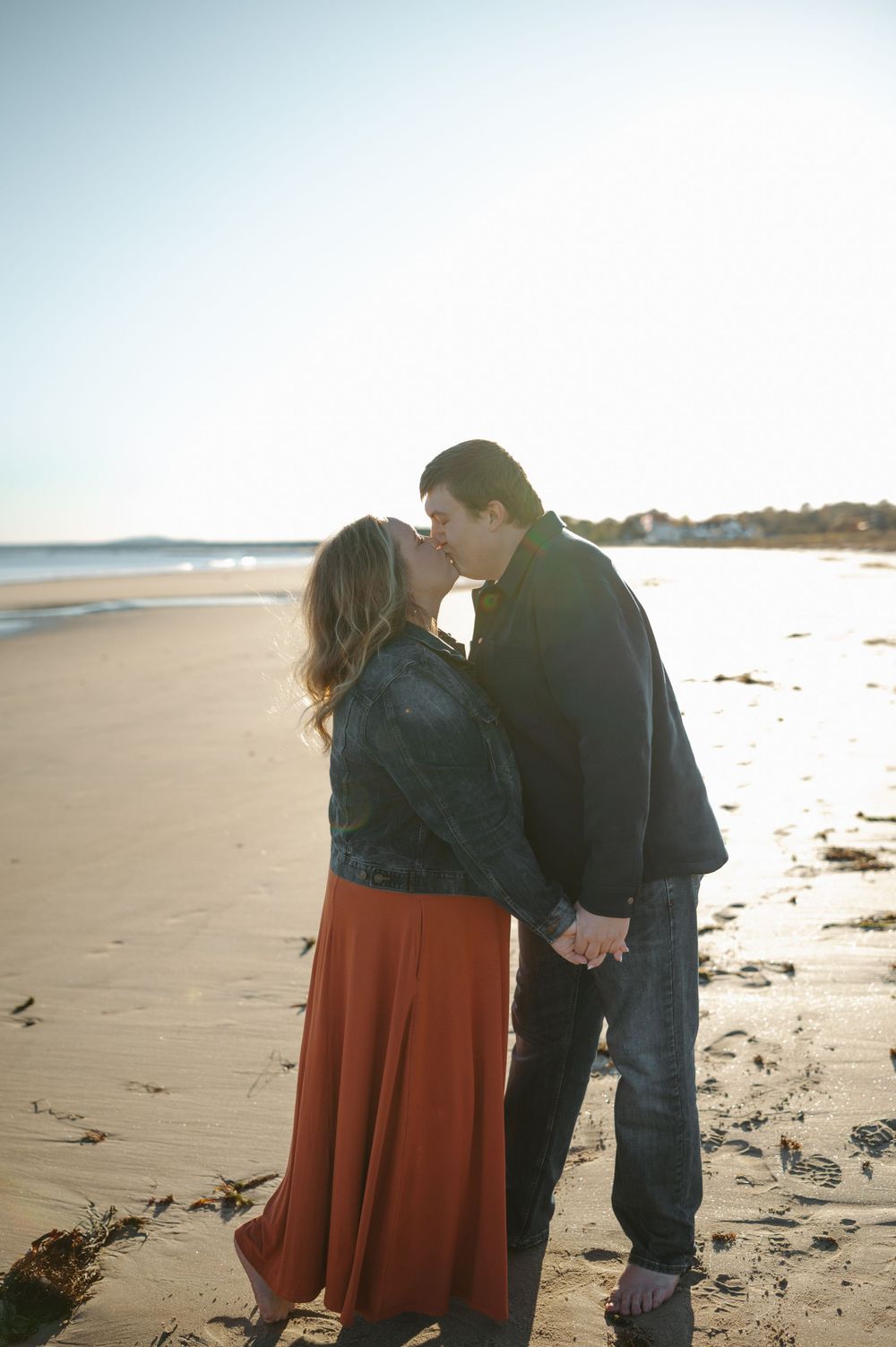 A couple walks together on a sunny beach during golden hour, casting long shadows on the sand.