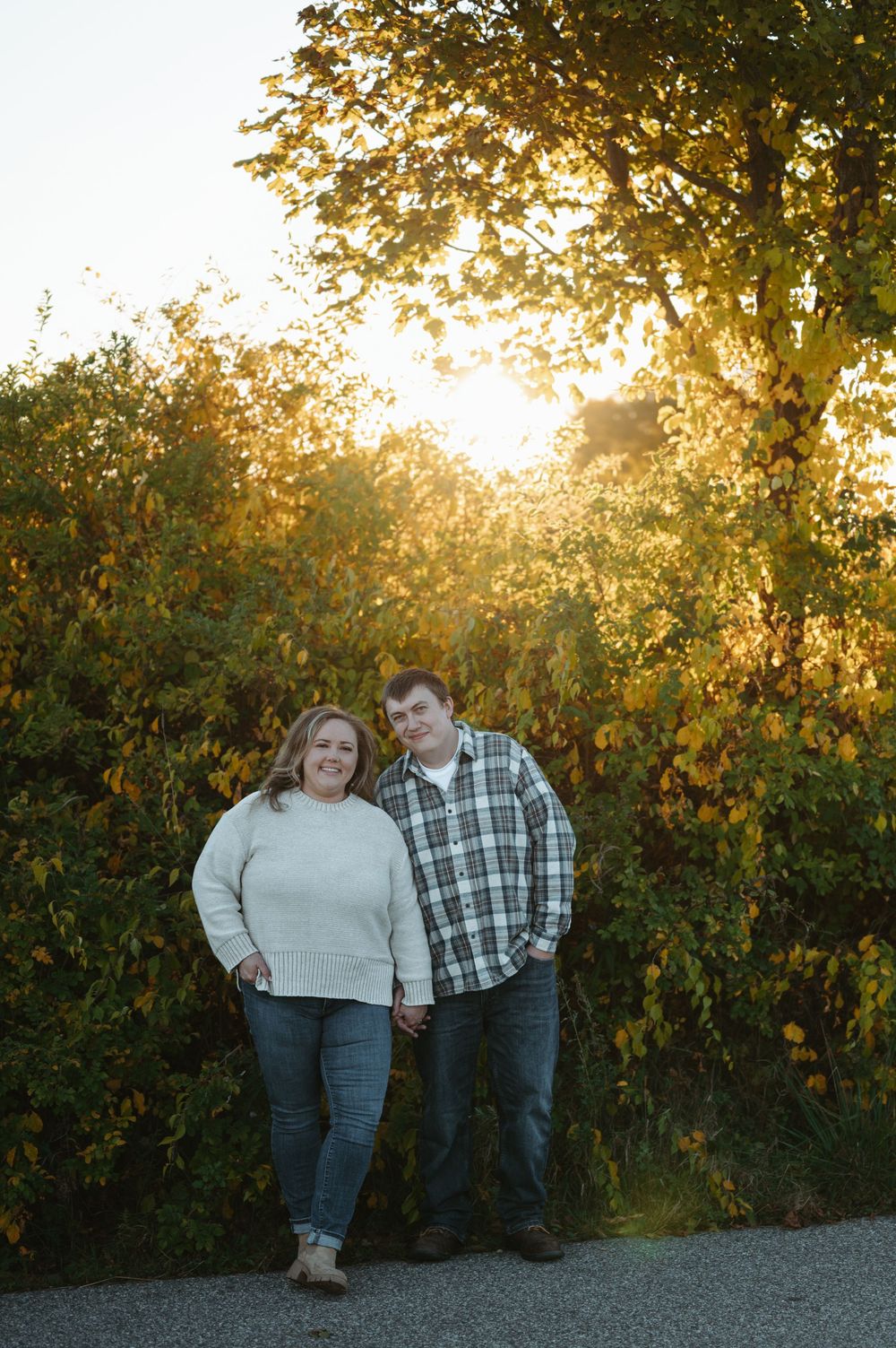 A couple poses together outdoors during golden hour with autumn foliage in the background.