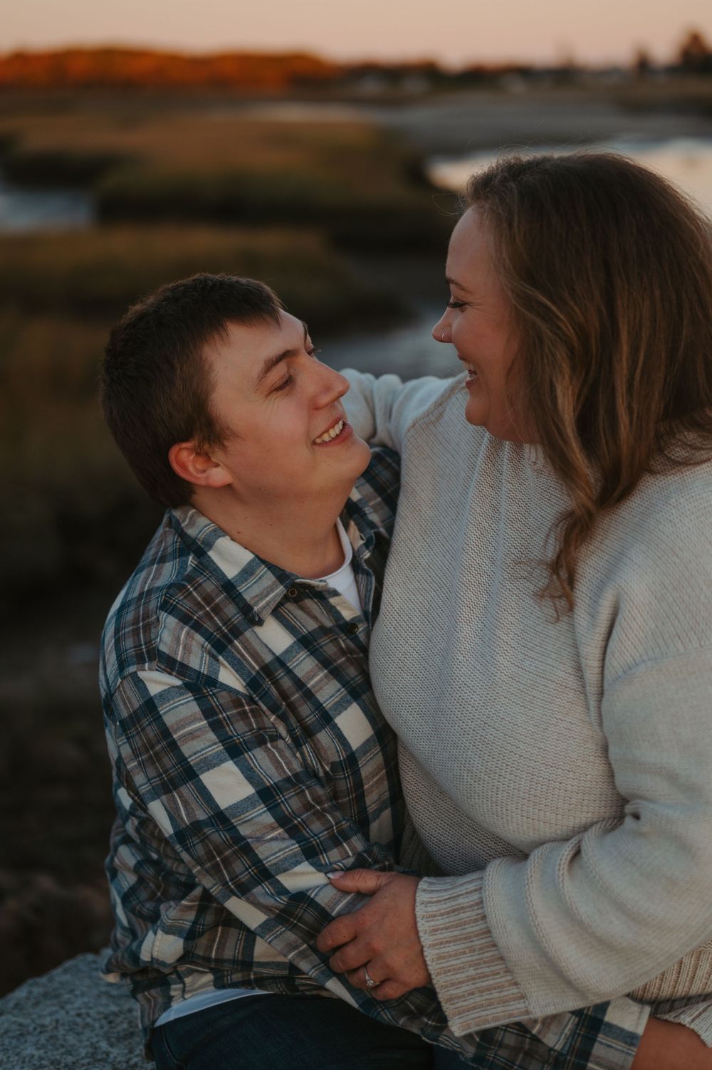 A mother and young son share playful laughter and cuddles while wearing cozy sweaters at sunset.