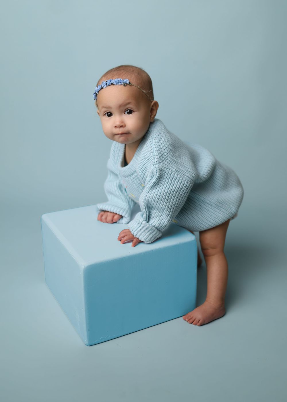 Baby in light blue sweater crawls on pale blue cube backdrop during minimalist portrait session.