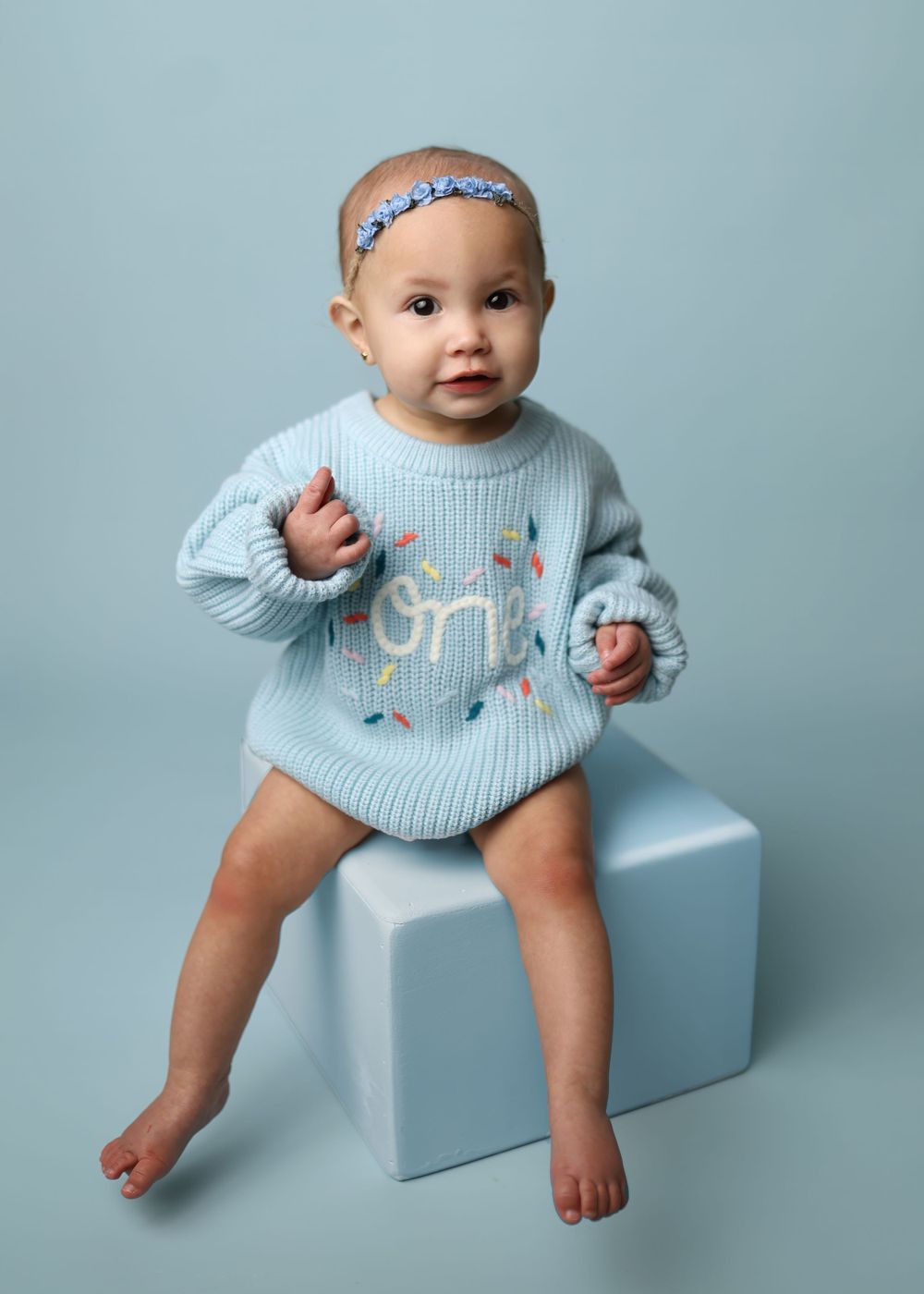 Baby wearing floral headband and embroidered sweater sits on light blue cube during studio photoshoot.