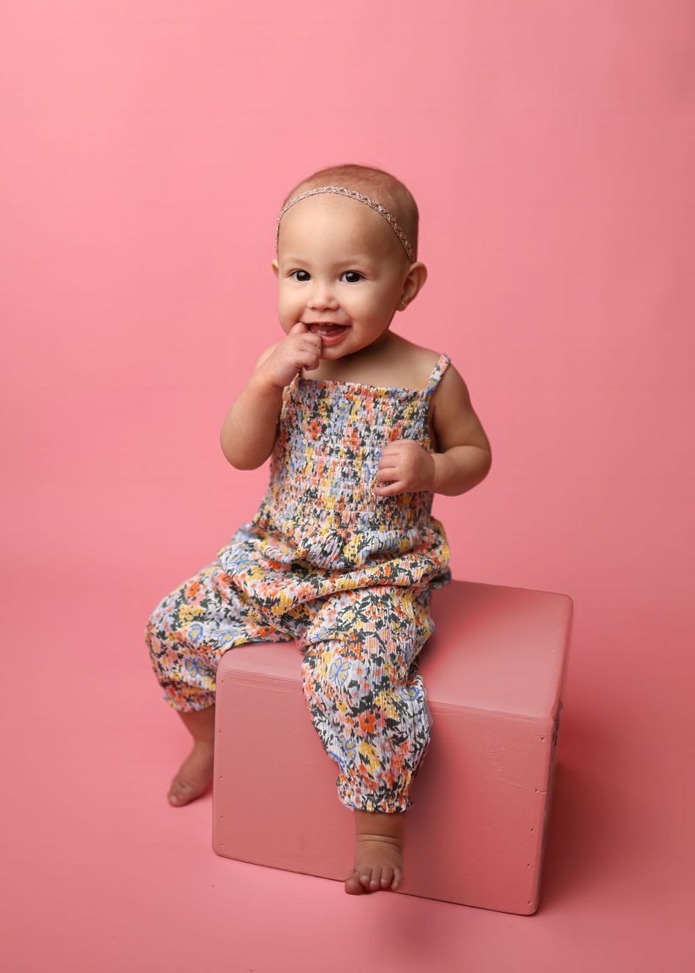 A baby in a floral print jumpsuit sits on a pink cube against a pink background while putting fingers in mouth.