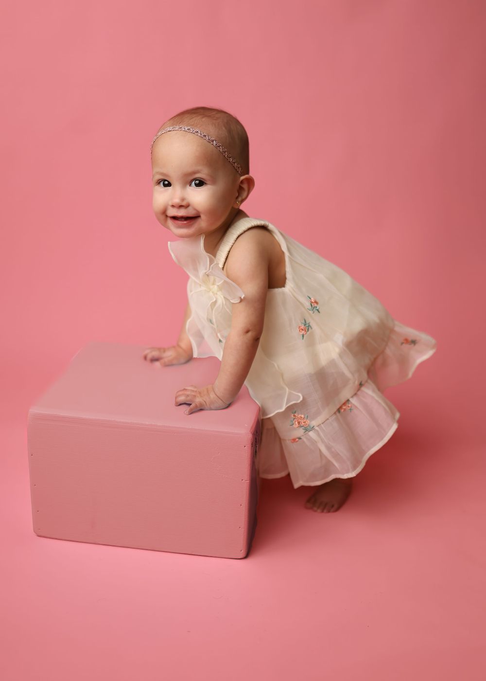 Small child in flowy cream dress leans playfully against pink cube against pink background during photo session.