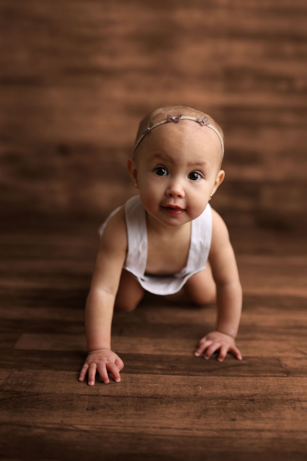A sweet baby in a white onesie crawls on a wooden floor while looking up at the camera with a bright expression.