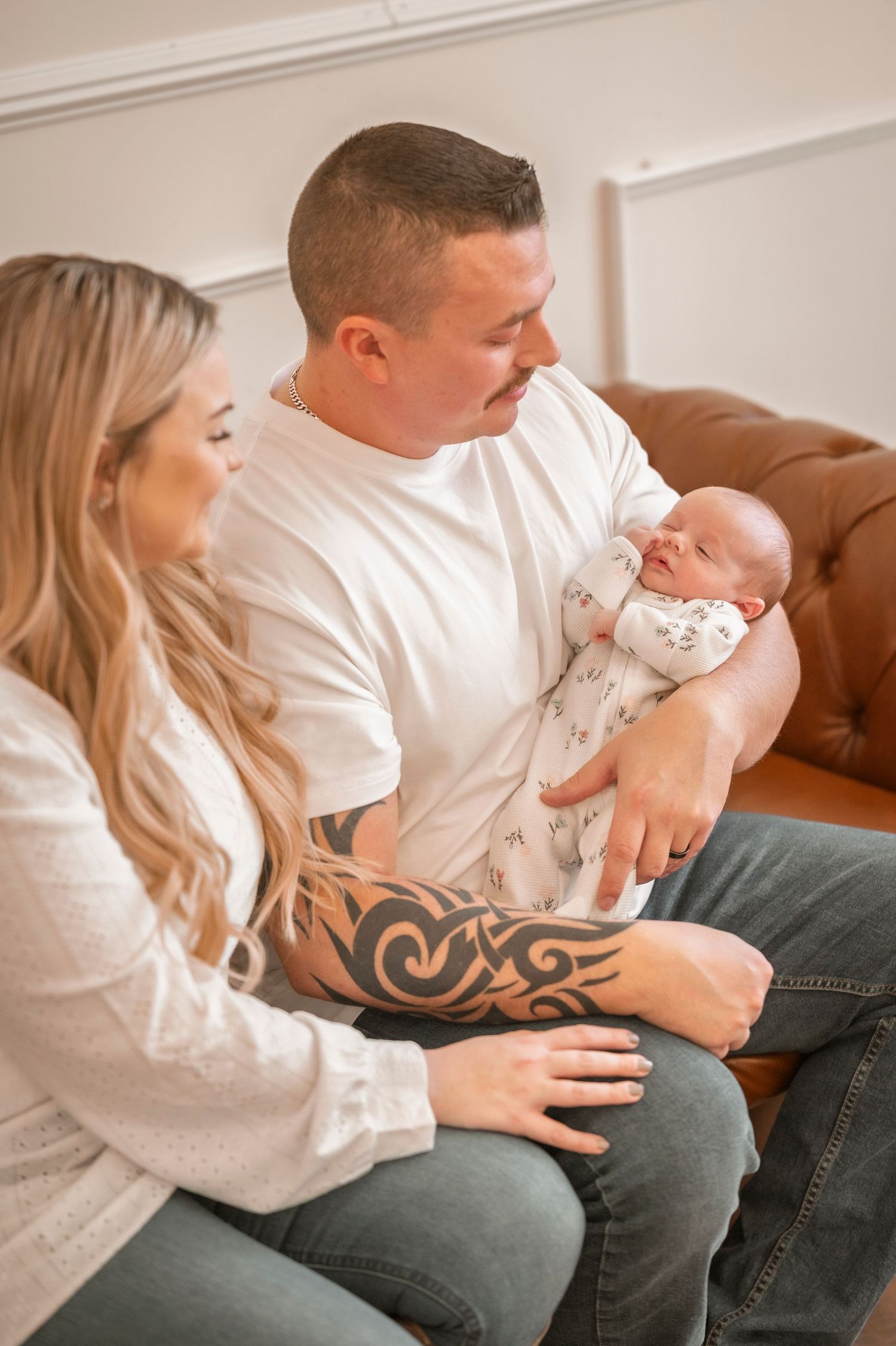Parents sitting together on leather couch holding and admiring their newborn baby, dad has tribal arm tattoo.