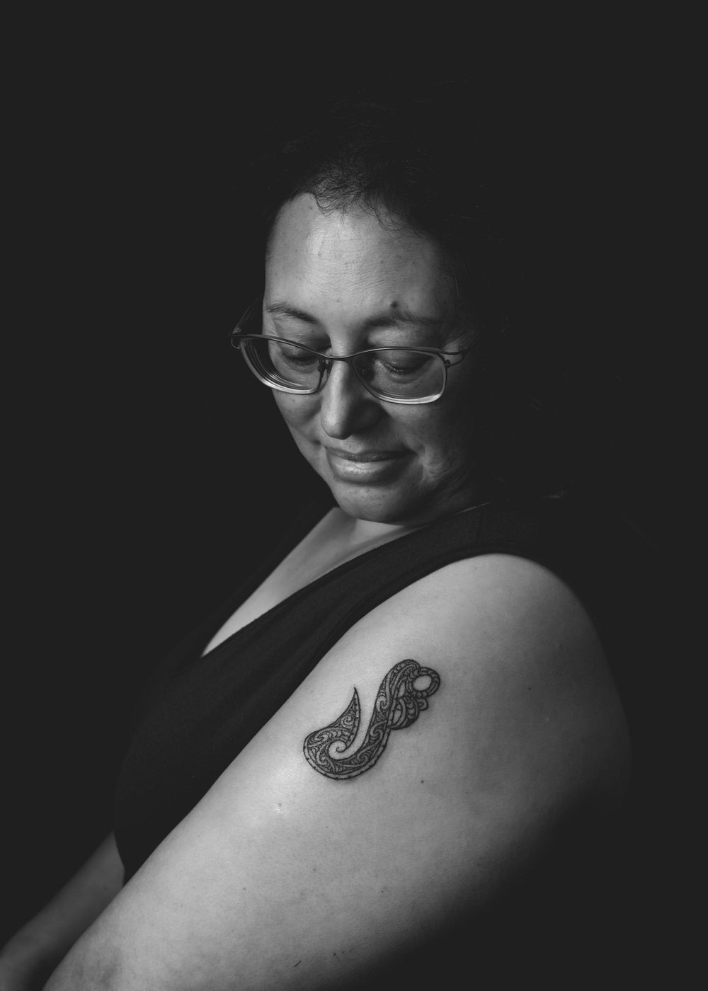 Black and white portrait of a person wearing glasses, with an elegant paisley tattoo visible on their bare shoulder.
