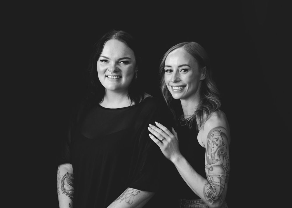 Black and white studio portrait of two people in black tops smiling together with visible tattoos on their arms.