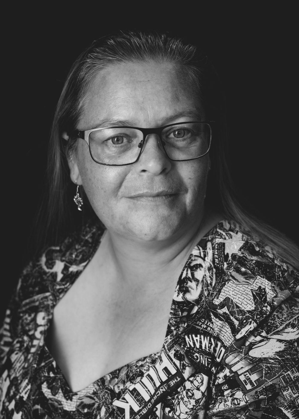 A black and white portrait of someone wearing glasses and a patterned top, shown against a dark background.