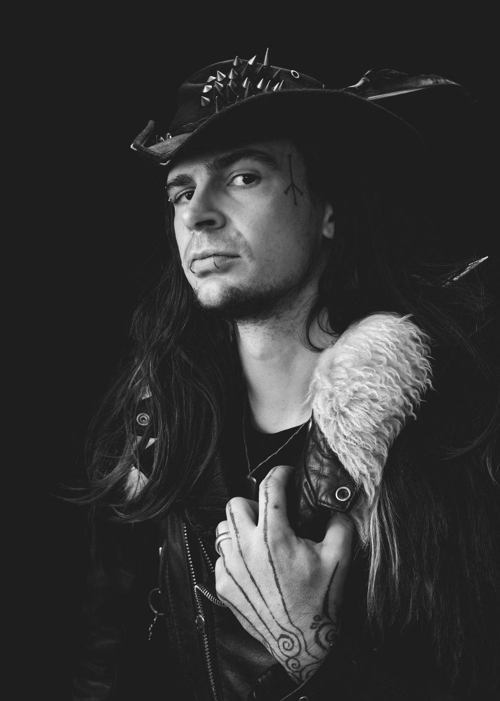 Dramatic black and white portrait of person in leather jacket with fur collar and cowboy hat with long dark hair.