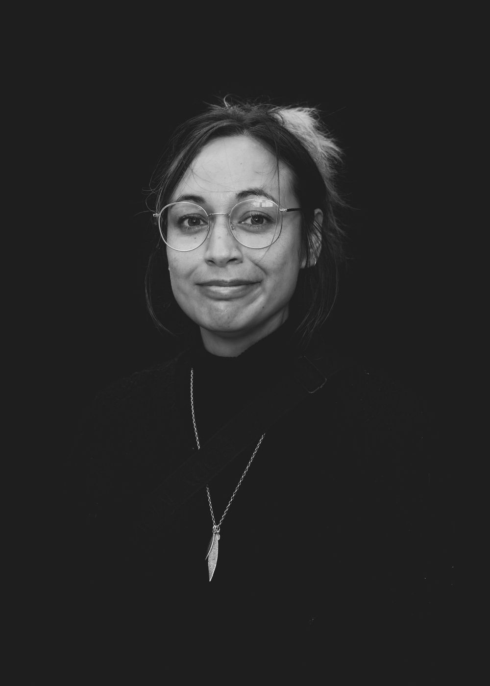 A black and white portrait of a person wearing round glasses and a necklace with a feather pendant, smiling warmly.