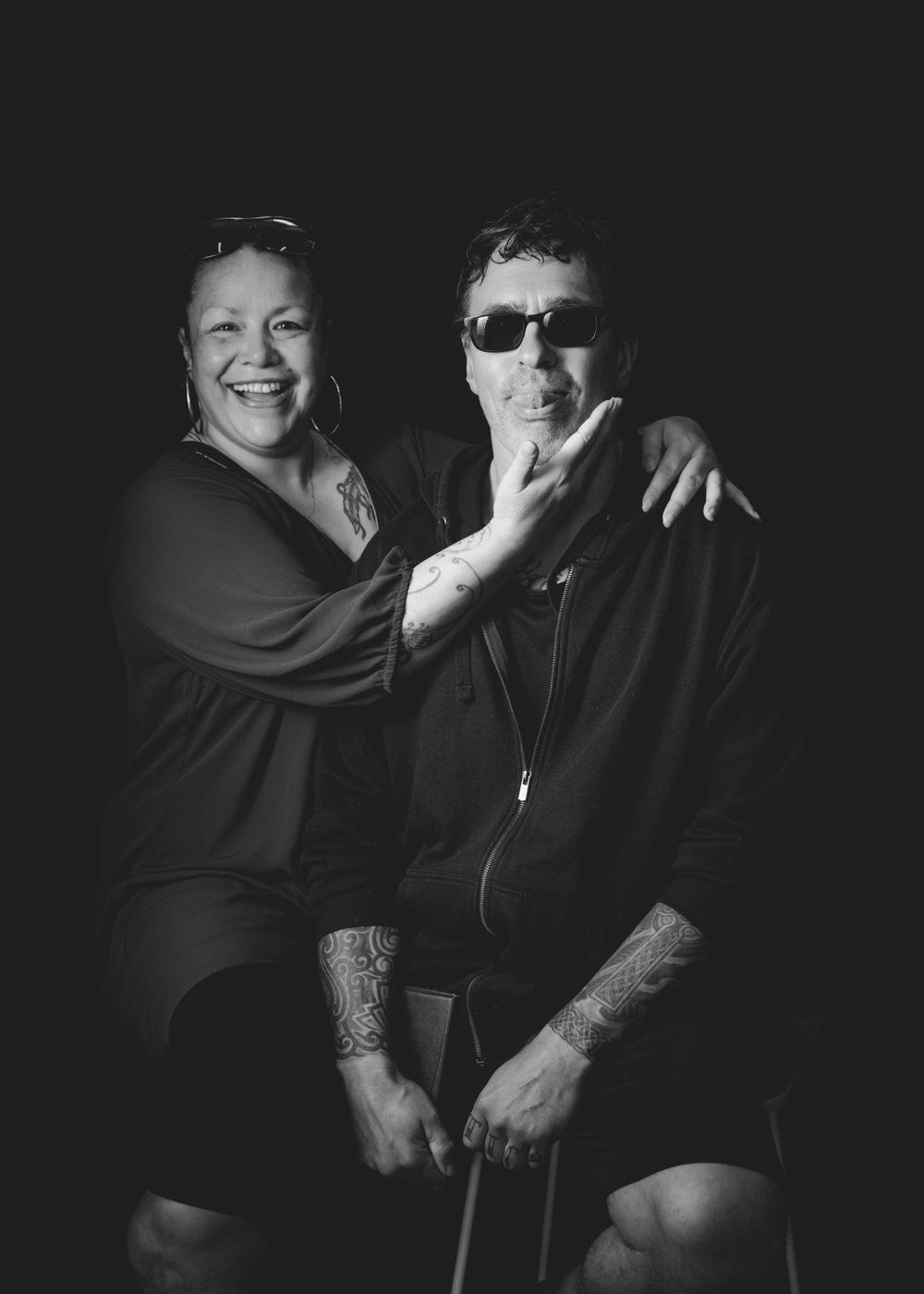 Black and white portrait photograph of two people embracing and smiling against a dark background.