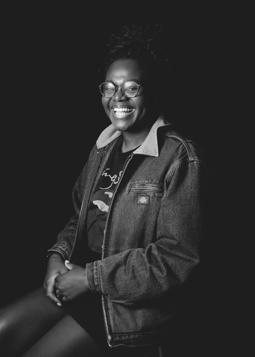 Black and white portrait of someone wearing glasses and a denim jacket, smiling joyfully against a dark background.