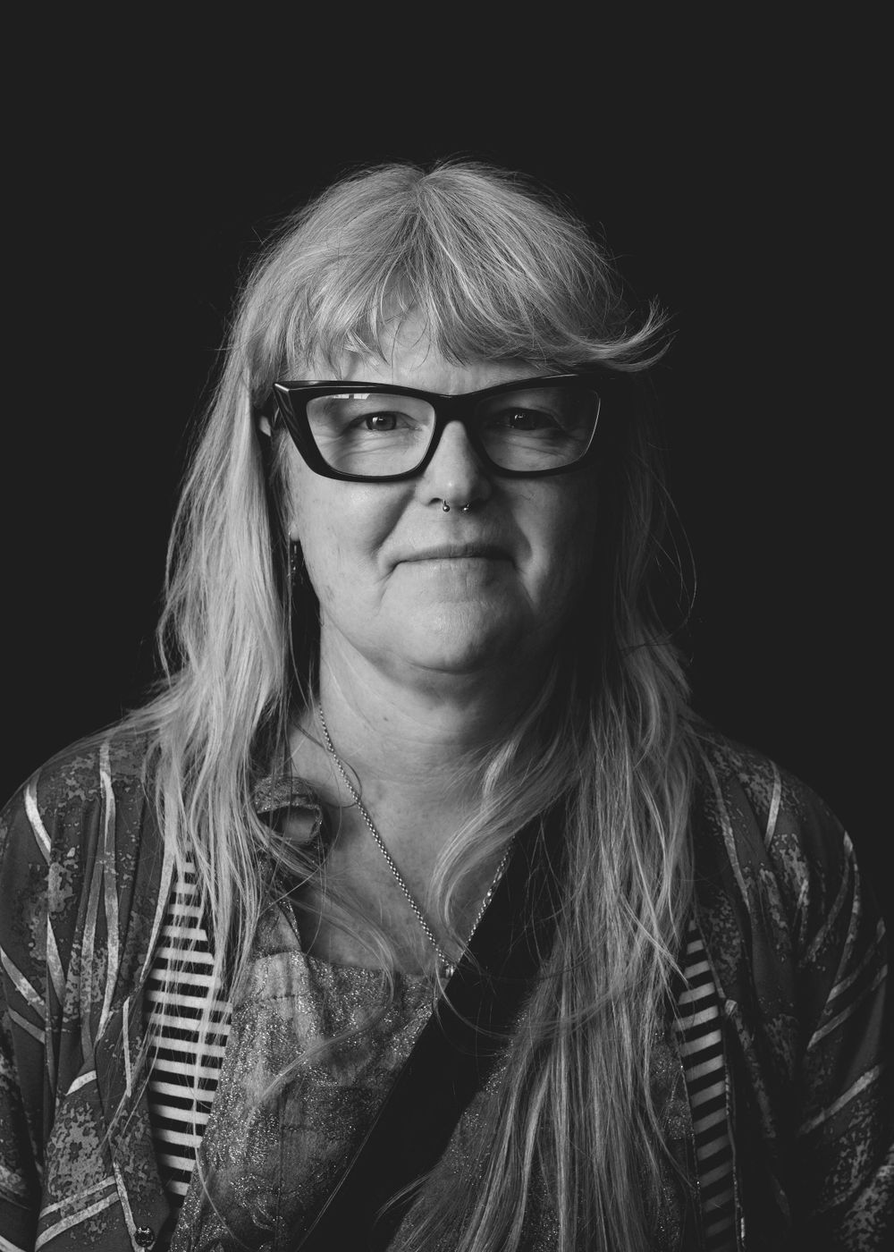 Black and white portrait of a person with long gray hair and glasses wearing a patterned top against dark background.