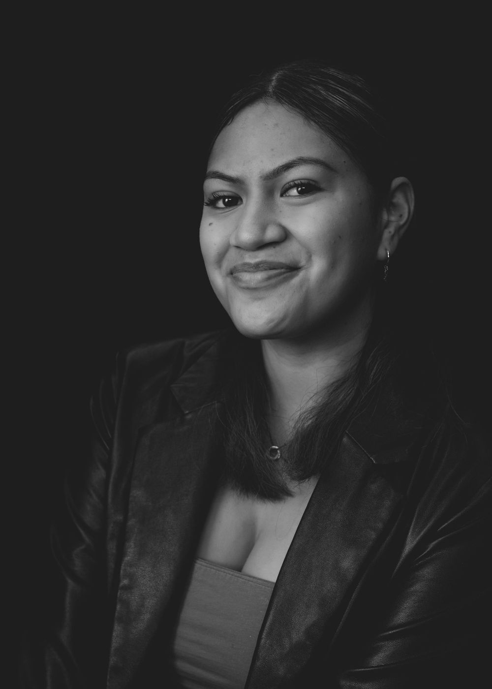 Black and white professional portrait photo of someone in a leather jacket smiling warmly against a dark background.