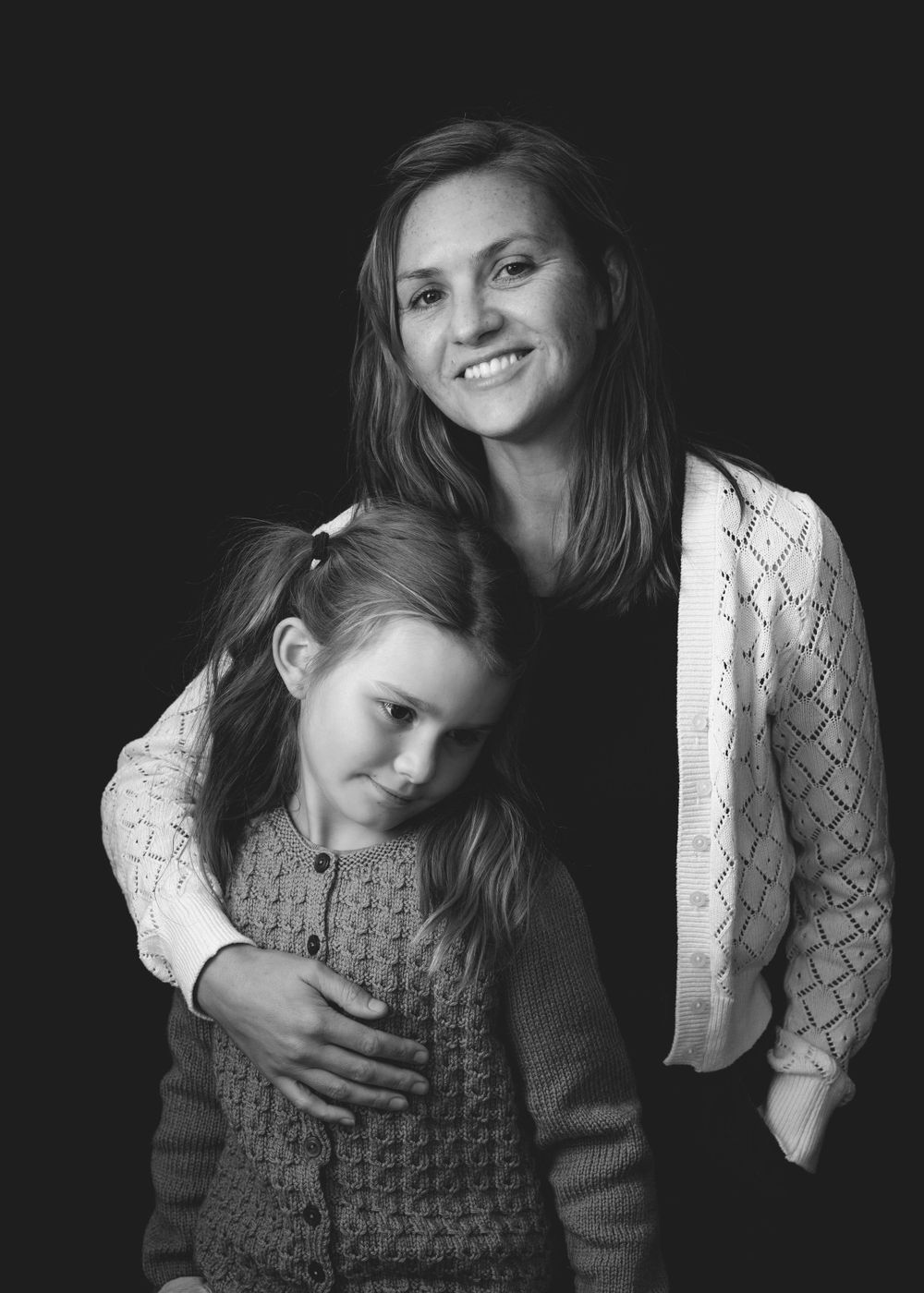 Black and white studio portrait of parent and child in knit sweaters embracing against dark background.