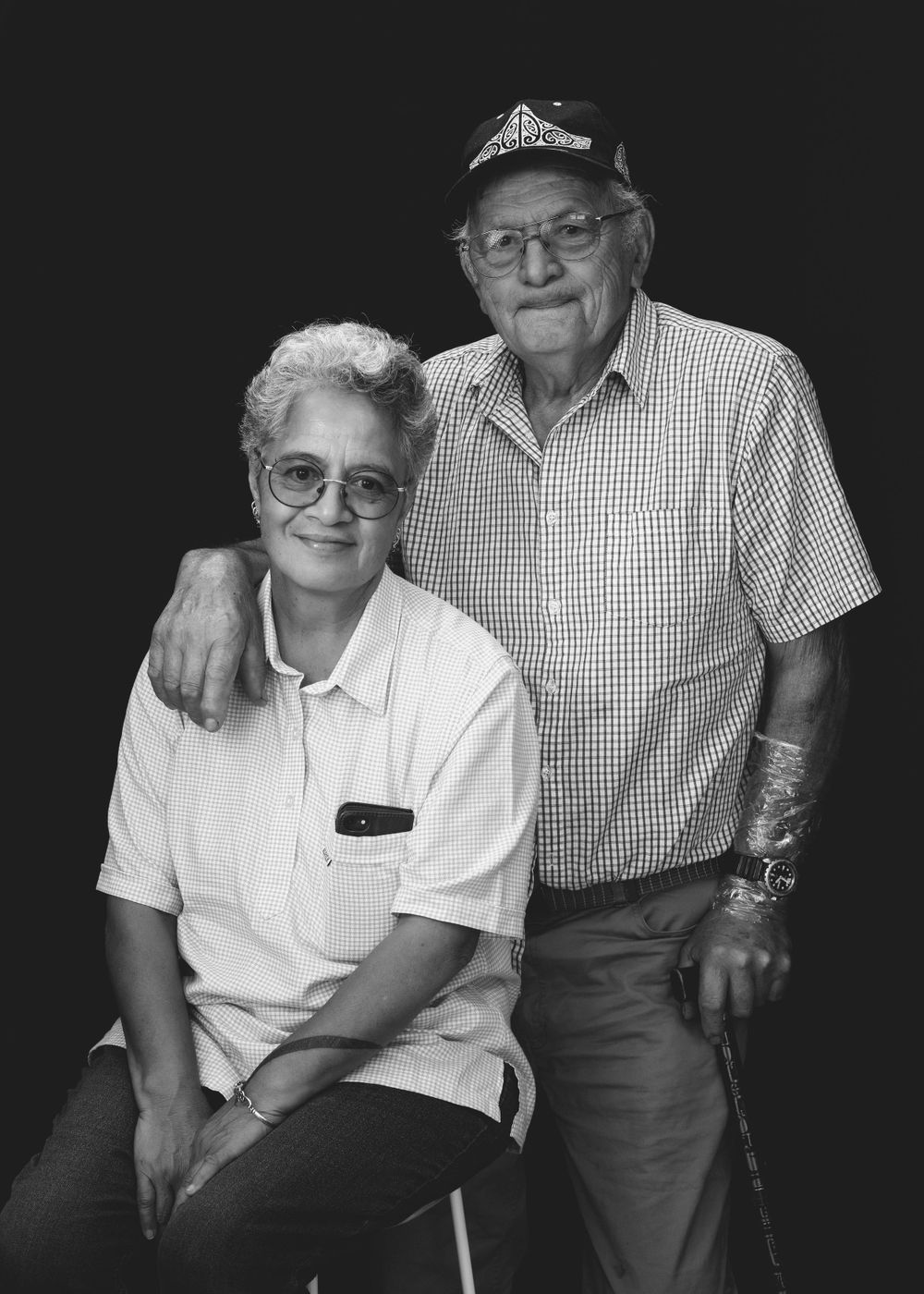 Black and white portrait of an elderly couple in casual attire sitting and standing together against a dark background.
