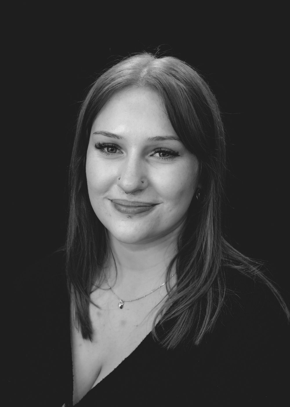 Black and white portrait of a smiling woman with straight dark hair and a delicate necklace against a dark background.