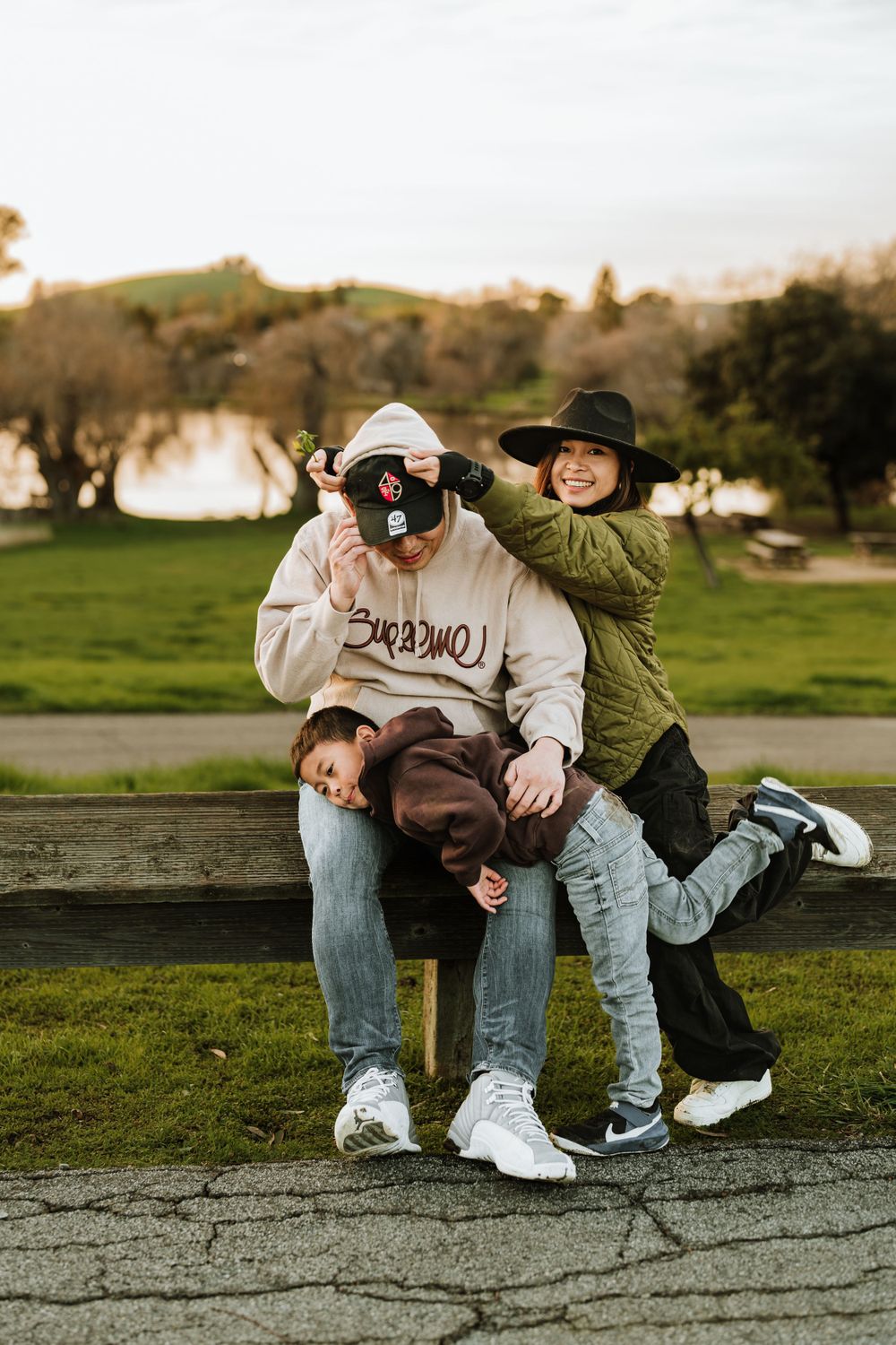 Two people playfully pose for a photo on a bench in a park at sunset with trees and a lake in the background.