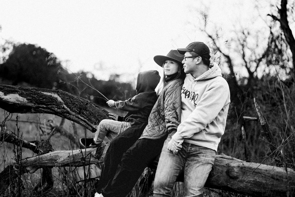 A black and white outdoor photograph of three people sitting together on a fallen log in a rural setting.