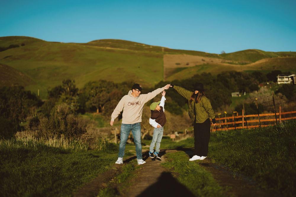 Three friends jump and dance together on a dirt path with rolling green hills and a blue sky in the background.