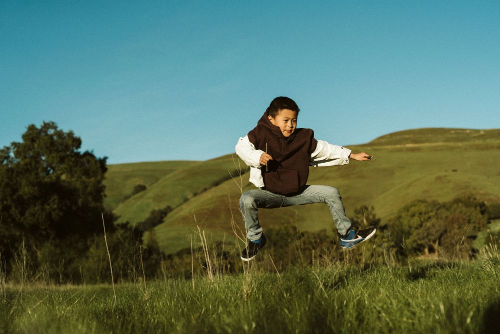 Person in brown jacket and jeans leaping energetically in a grassy field against blue sky on sunny day.