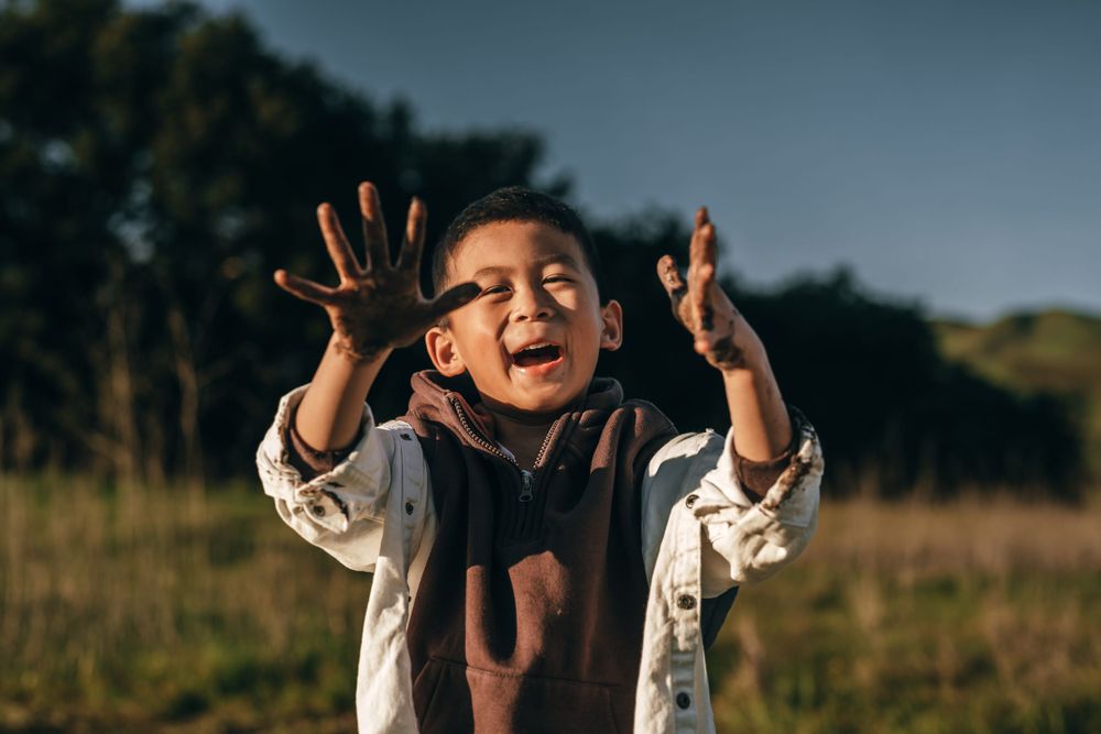 Child in brown shirt expressing pure joy with raised hands and laughing outdoors in golden sunlight.