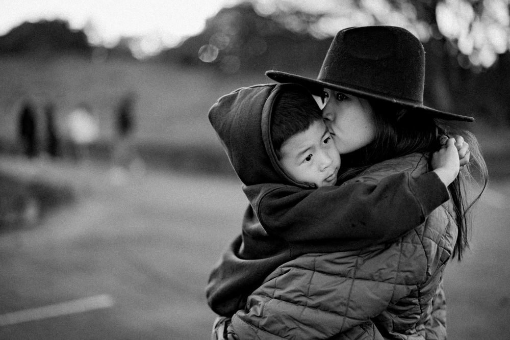 Black and white image showing a close embrace between two people wearing dark clothing outdoors with soft bokeh background.