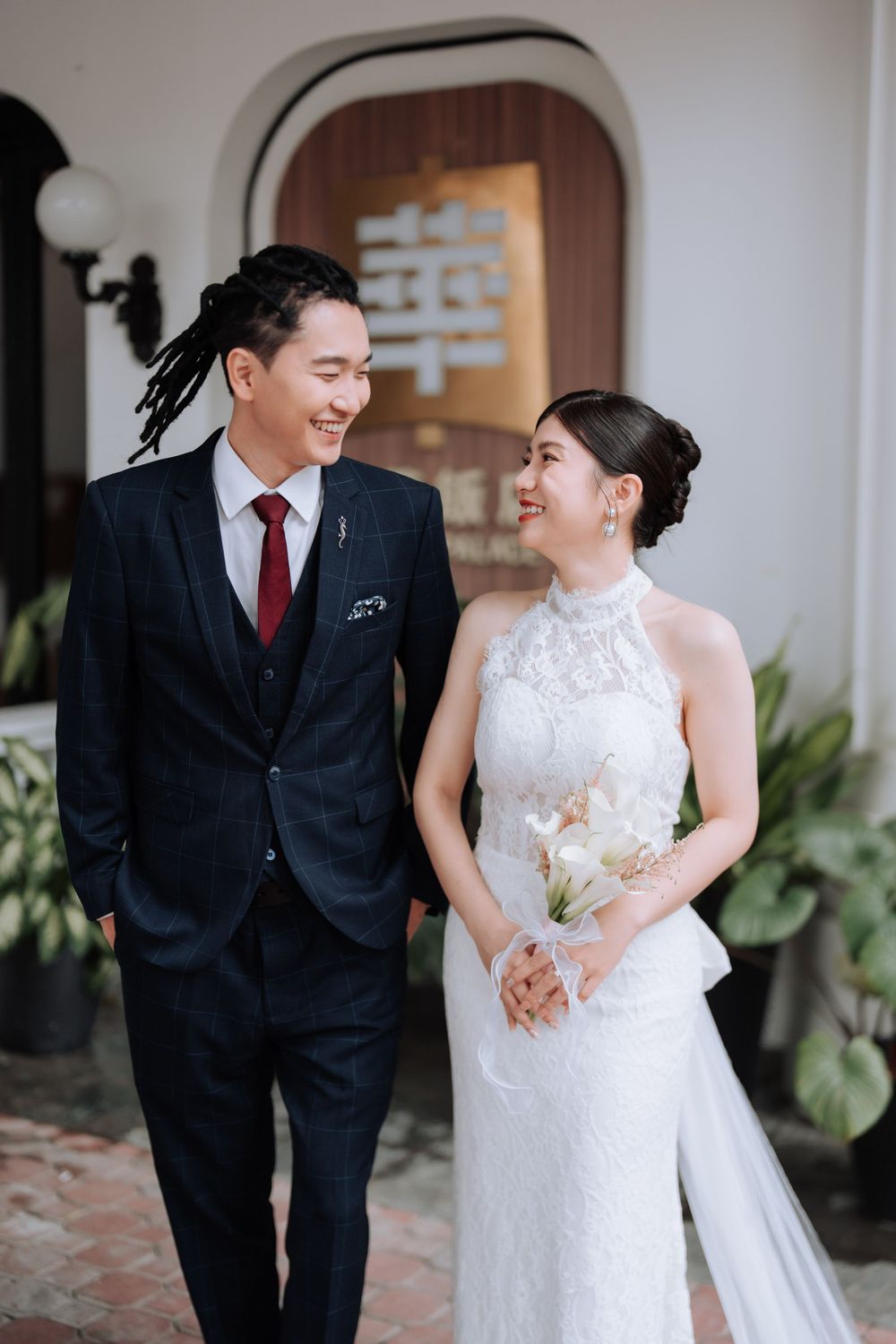 A wedding couple in traditional attire share a joyful moment against a white wall with Chinese characters.