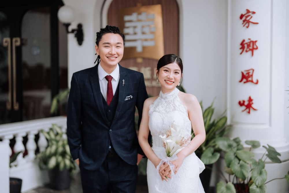 Newlyweds pose before a white building with Chinese characters wearing formal wedding attire.