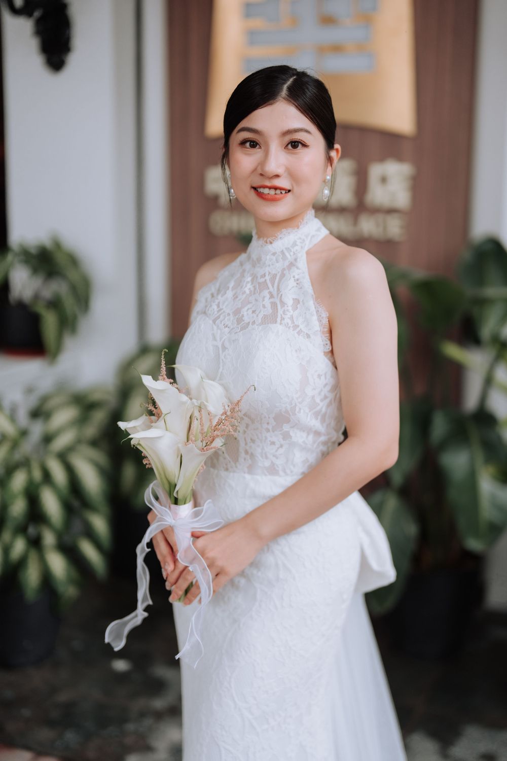 A bride in a white halter gown holds lilies while smiling beside potted plants.