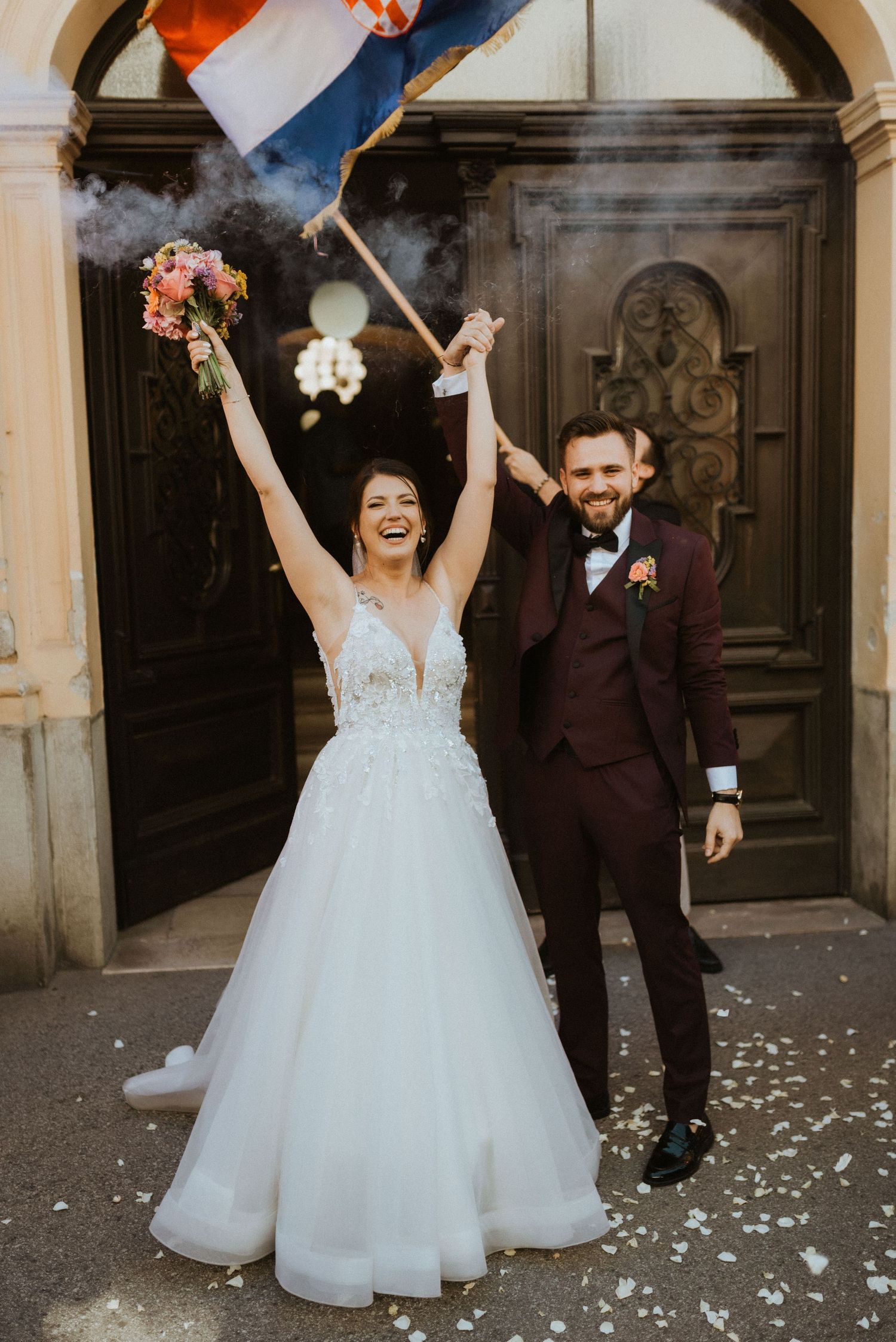 Joyful newlyweds celebrate outside historic doorway with a flag and bouquet amidst confetti and smoke.