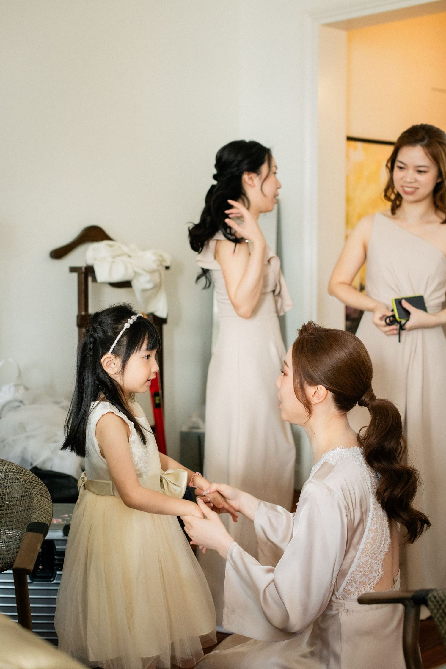 A bride in a white robe kneels to interact with a young flower girl while bridesmaids prepare in the background.
