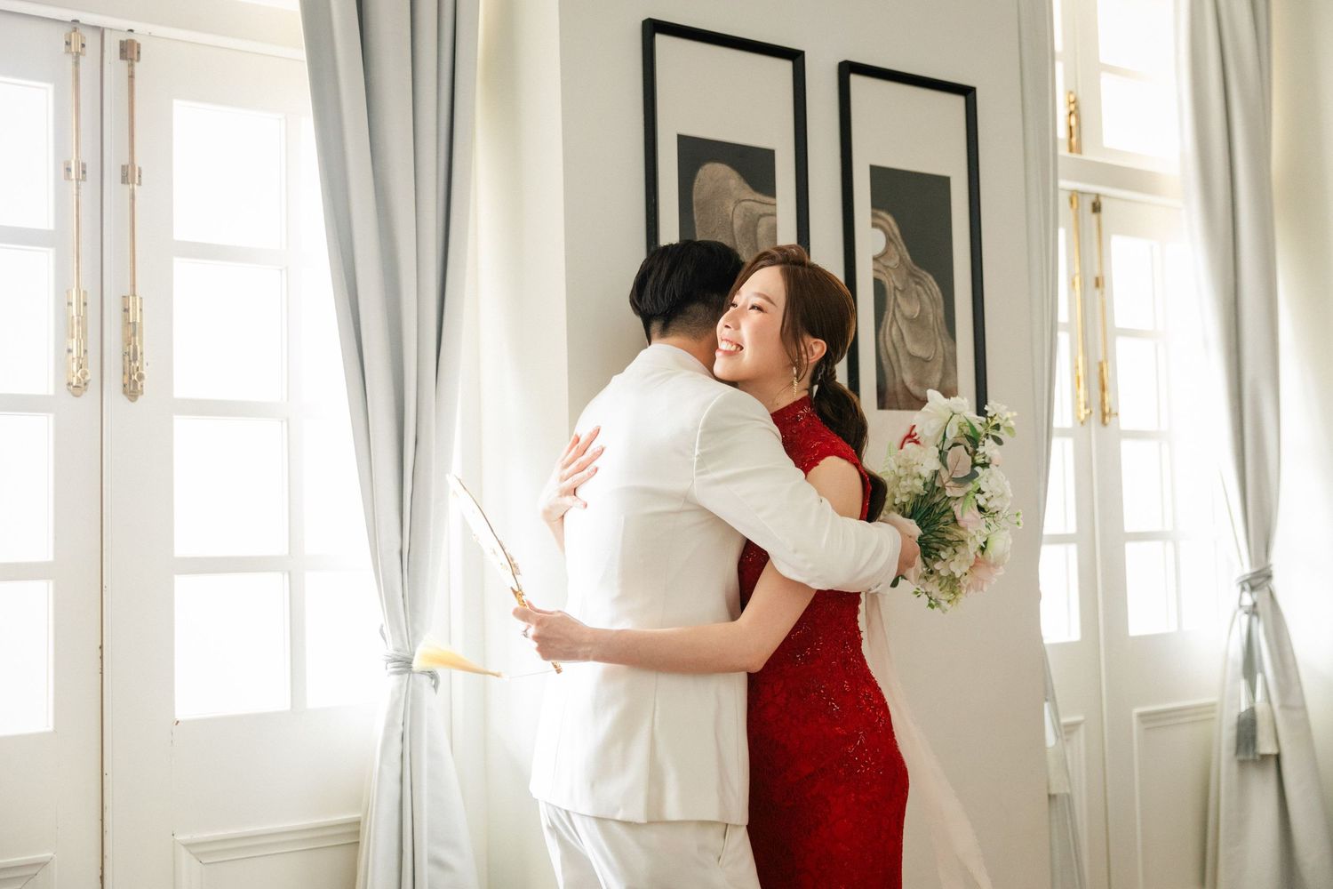 Bride in red qipao holding bouquet shares joyful embrace with groom in white suit by window.