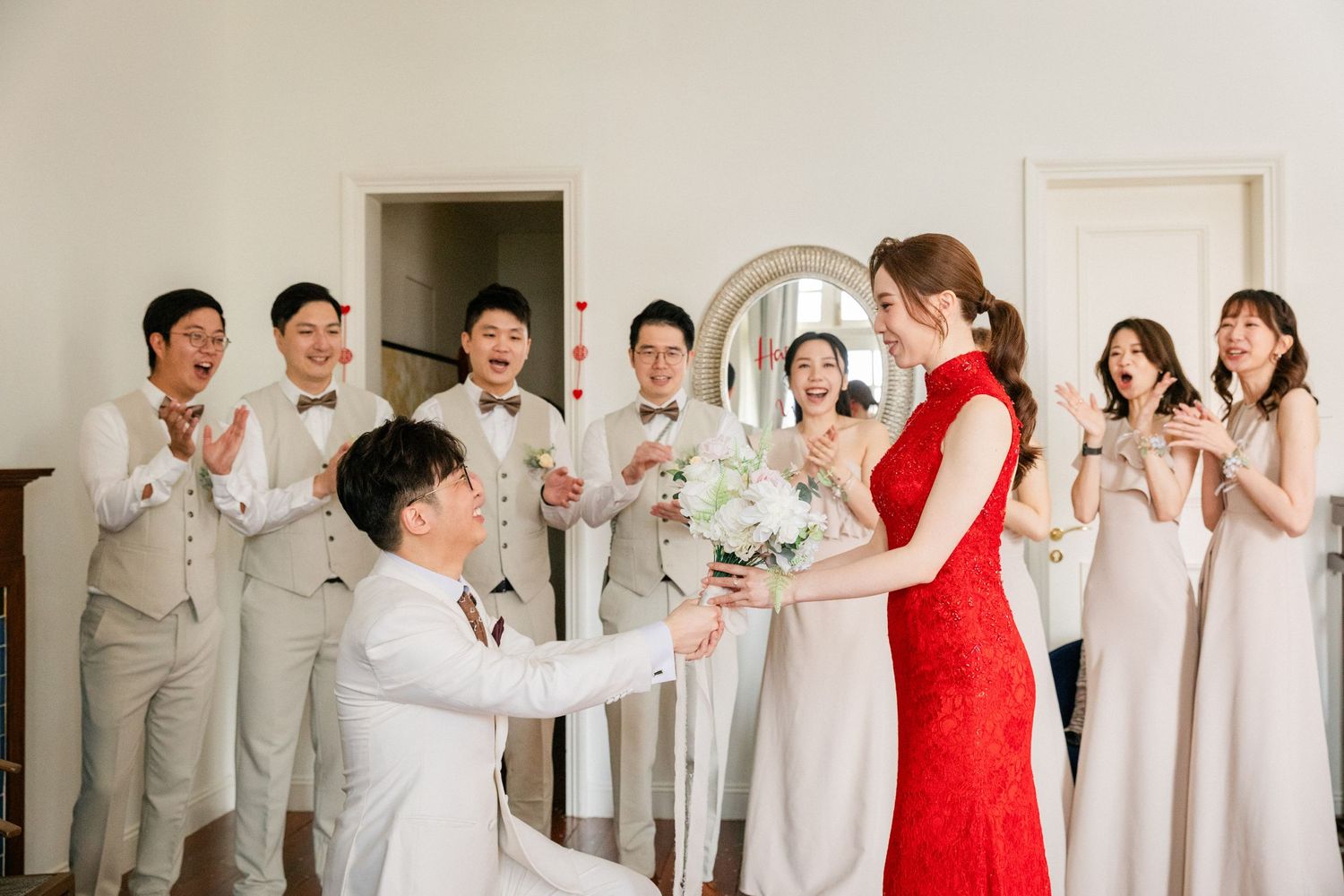 Groom kneels presenting bouquet to bride in red qipao surrounded by cheering wedding party.